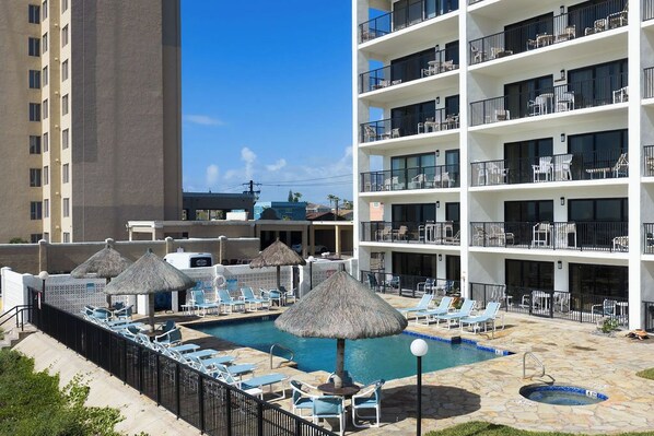 Un hotel con área de piscina, incluyendo sillones reclinables y sombrillas, y un edificio de varios pisos con balcones.