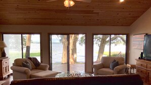 Un salón amplio con un sofá grande, dos sillones y una mesa de centro. Hay ventanas grandes que ofrecen vistas a un lago y árboles.