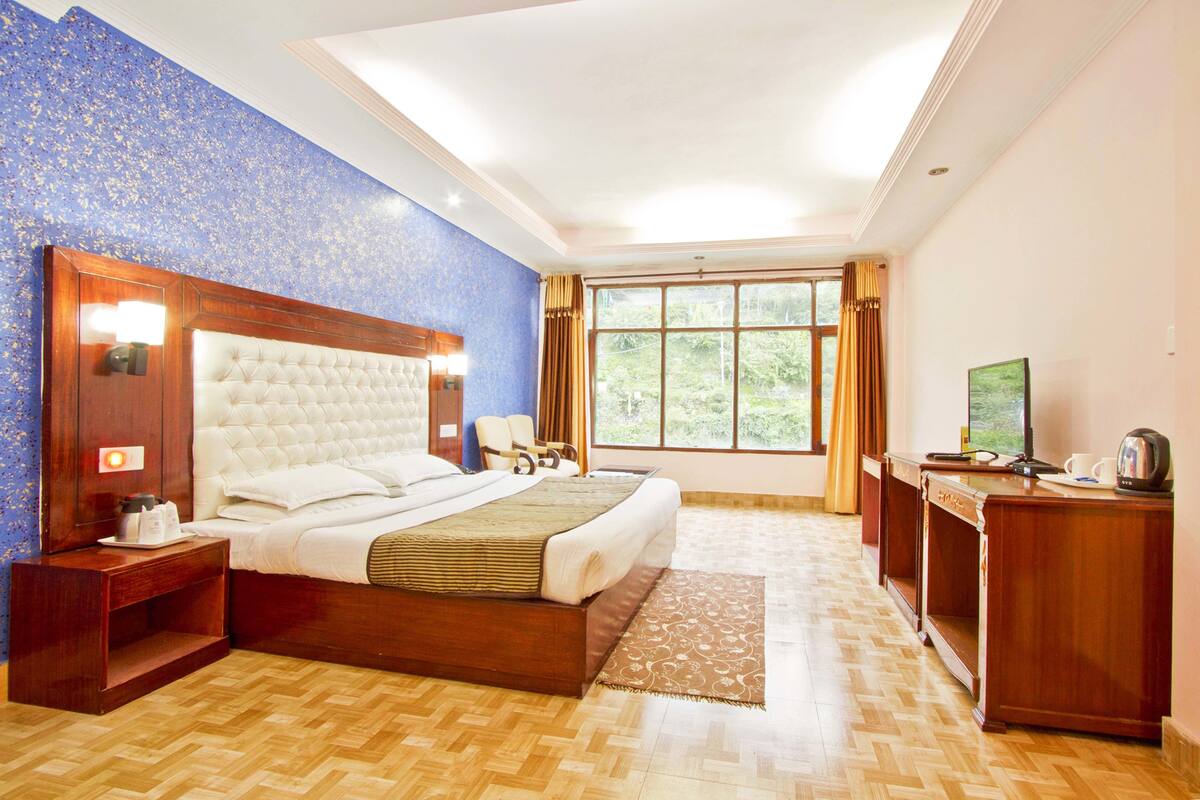 Ein Hotelzimmer mit einem großen Bett, einem Holzkopfteil, Nachttischen, einem Schreibtisch und Blick ins Grüne durch das Fenster.