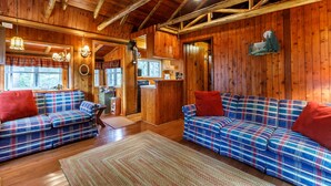 L’intérieur d’une cabane est caractérisé par des murs et des plafonds en bois, des canapés à carreaux avec des coussins rouges et un coin cuisine.