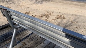 Une terrasse en bois avec une rambarde en métal, donnant sur une plage de sable et une mer calme.