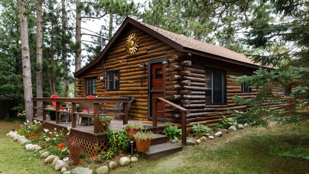 Une cabane en rondins dotée d’une terrasse, entourée d’un jardin agrémenté de rochers et de plantes.