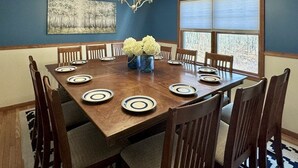Een eetzaal met een houten tafel voor zes personen, omringd door stoelen, een kroonluchter en een raam met luxaflex.