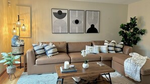 Een woonkamer met een bruine modulaire bank, een houten salontafel en een grote potplant. Boven de bank hangen abstracte kunstwerken.