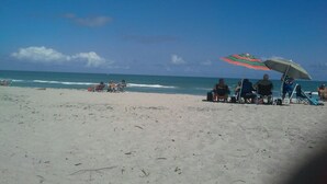 Ein Strand mit Menschen, die unter Sonnenschirmen sitzen, einem klaren Himmel und dem Ozean im Hintergrund.