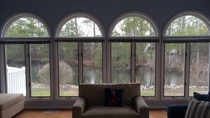 Un salon avec un fauteuil beige, une table basse en verre et une vue sur un lac, visible à travers de grandes fenêtres en arc de cercle.
