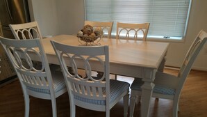 Une table à manger avec quatre chaises blanches, un panier en osier posé sur la table, et une fenêtre avec des stores en arrière-plan.