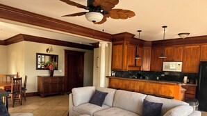 A living room with a sectional sofa, ottoman, wooden ceiling fan, and a kitchen with wooden cabinets and a microwave.