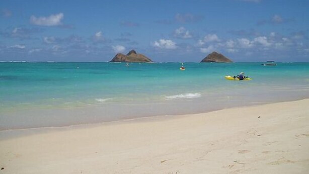 A beach with clear turquoise water, two small islands in the distance, and a person kayaking on the sand.