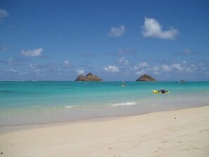 A beach with clear turquoise water, two small islands in the distance, and a person kayaking on the sand.