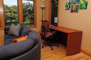 A modern living room with a grey sofa, a wooden desk, and a view of a green landscape through large windows.
