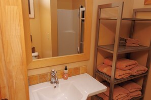 Un baño con un lavamanos blanco, un espejo, una repisa con toallas dobladas y una zona de ducha.