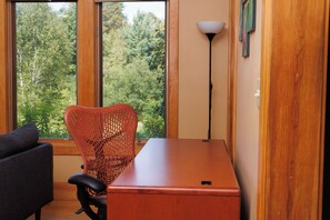 A room with a wooden desk, a wicker chair, a black sofa, and a view of trees through a large window.