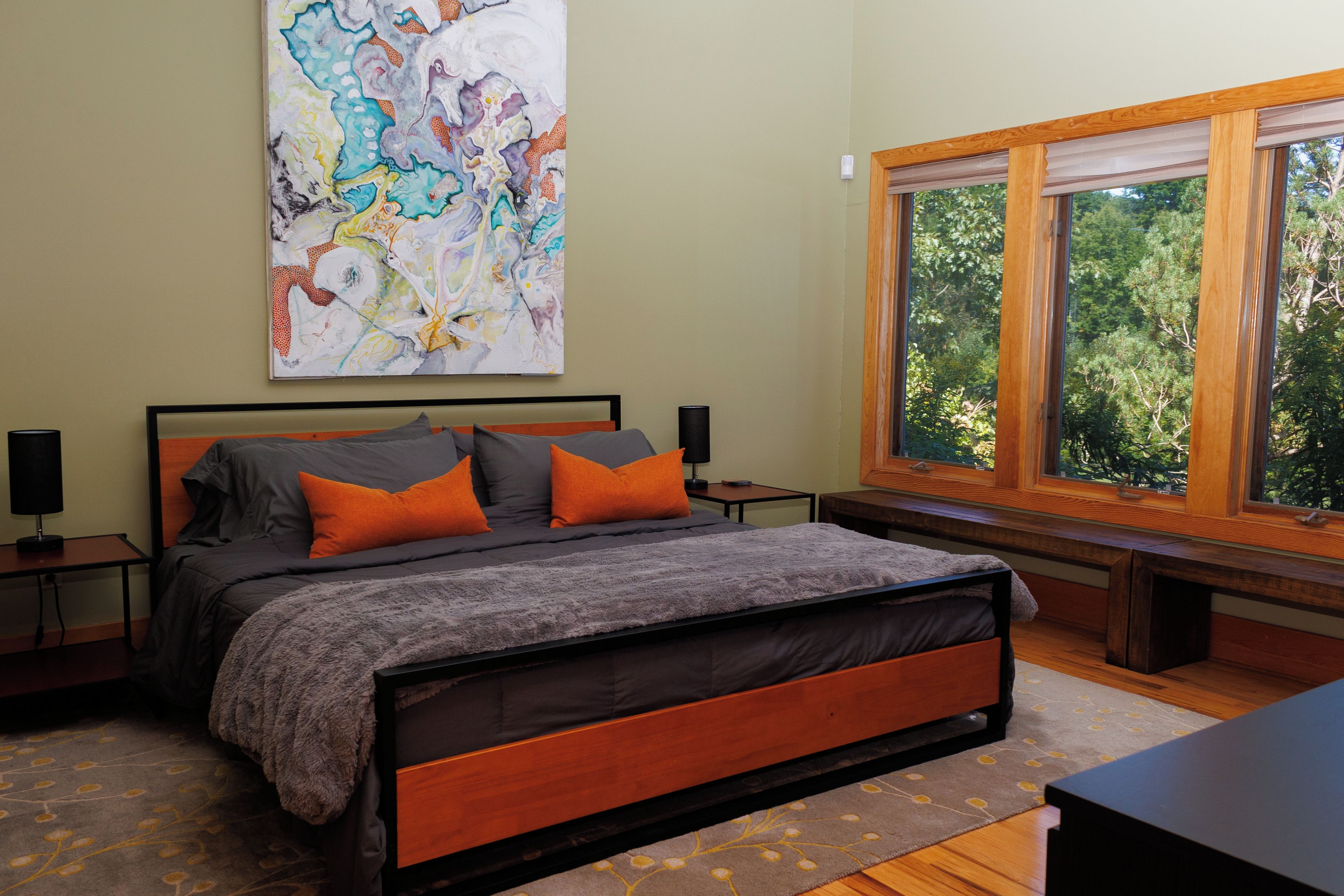 A bedroom with a bed, a nightstand, a lamp, a window with a view of trees, and a large abstract painting on the wall.