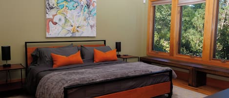 A bedroom with a bed, a nightstand, a lamp, a window with a view of trees, and a large abstract painting on the wall.