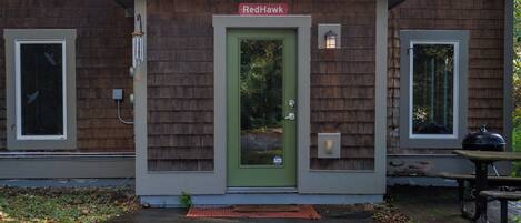A building with a green door and a sign above it that reads 'Red Hawk.' There are two windows on either side of the door, and a small outdoor table is visible to the right.