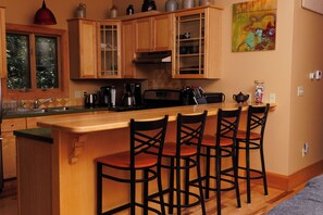 Une cuisine avec des meubles de rangement en bois, une machine à café, une bouilloire et une cuisinière. On y trouve quatre tabourets de bar avec des pieds noirs.
