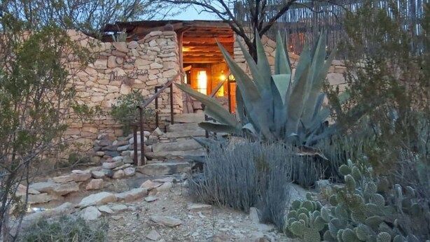 A stone building with a wooden staircase leading to a door, surrounded by cacti and desert vegetation.