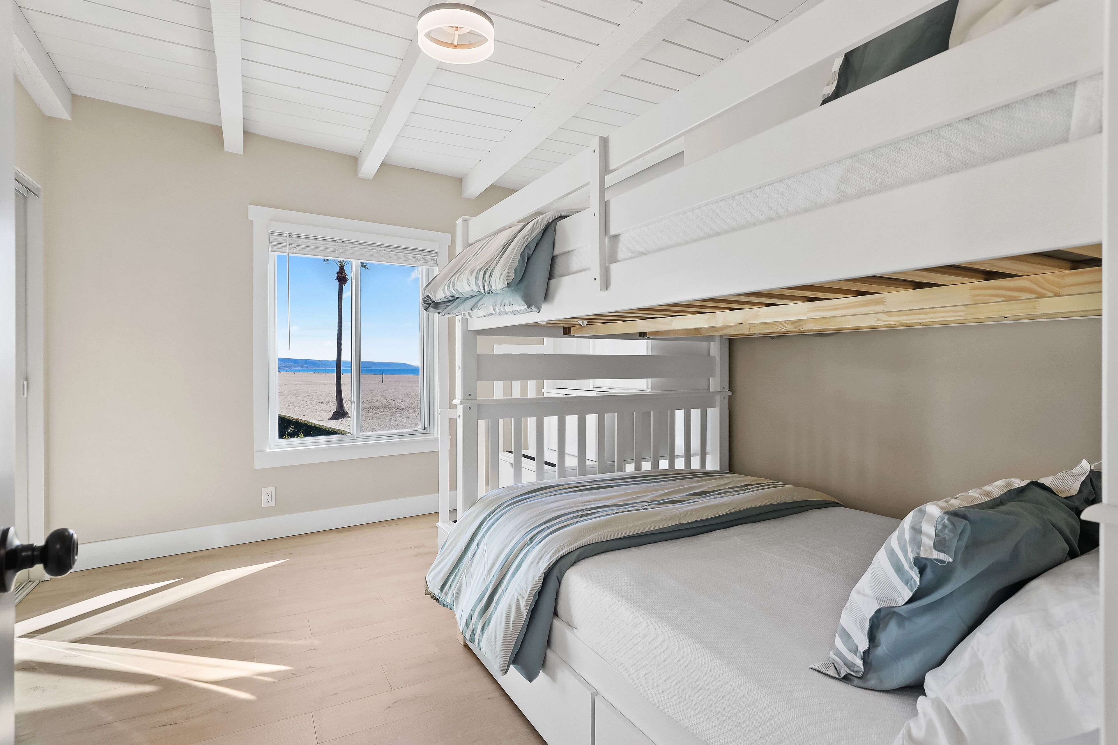 A bunk bed room with a window overlooking a beach, a ceiling fan, and a wooden floor.