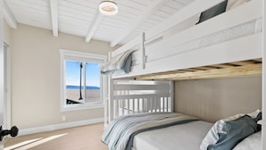 Une chambre avec un lit superposé, une fenêtre donnant sur la plage, un ventilateur de plafond et un plancher en bois.