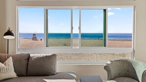 Une pièce dotée d’une grande fenêtre donnant sur une plage, d’un canapé, d’un fauteuil et d’une lampe.