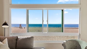 A room with a large window overlooking a beach, a sofa, a chair, and a lamp.