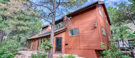 A two-story wooden cabin with a stone foundation, surrounded by pine trees and a wooden deck.