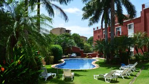 A swimming pool surrounded by lush greenery and palm trees, with lounge chairs and a building in the background.
