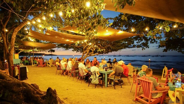 An outdoor dining area with tables and chairs under a canopy, overlooking a beach with boats in the distance.