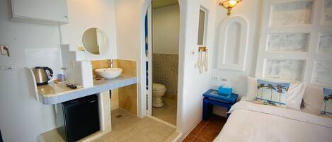 A bathroom with a sink, mirror, and toilet. There is a shower area with a tub and a small table with a towel and a box.