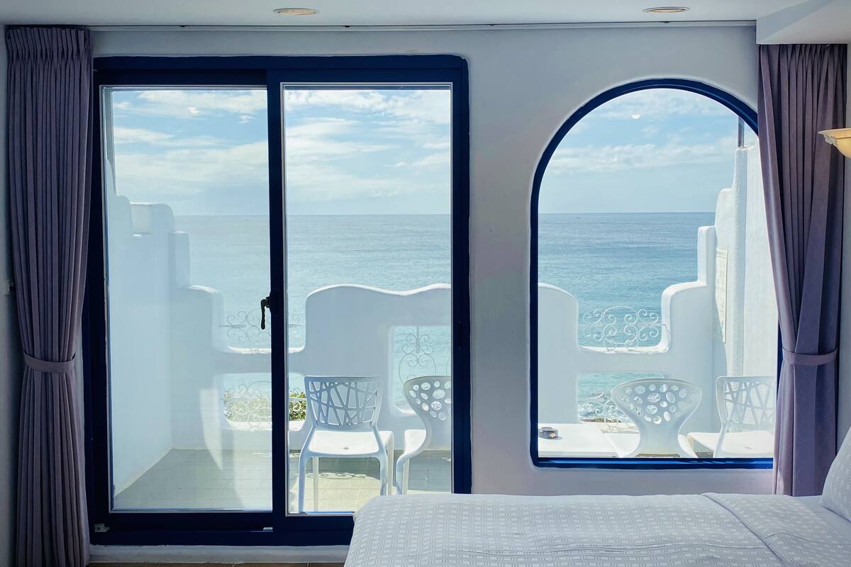A bedroom with a large sliding glass door leading to a balcony with white chairs and a view of the sea.