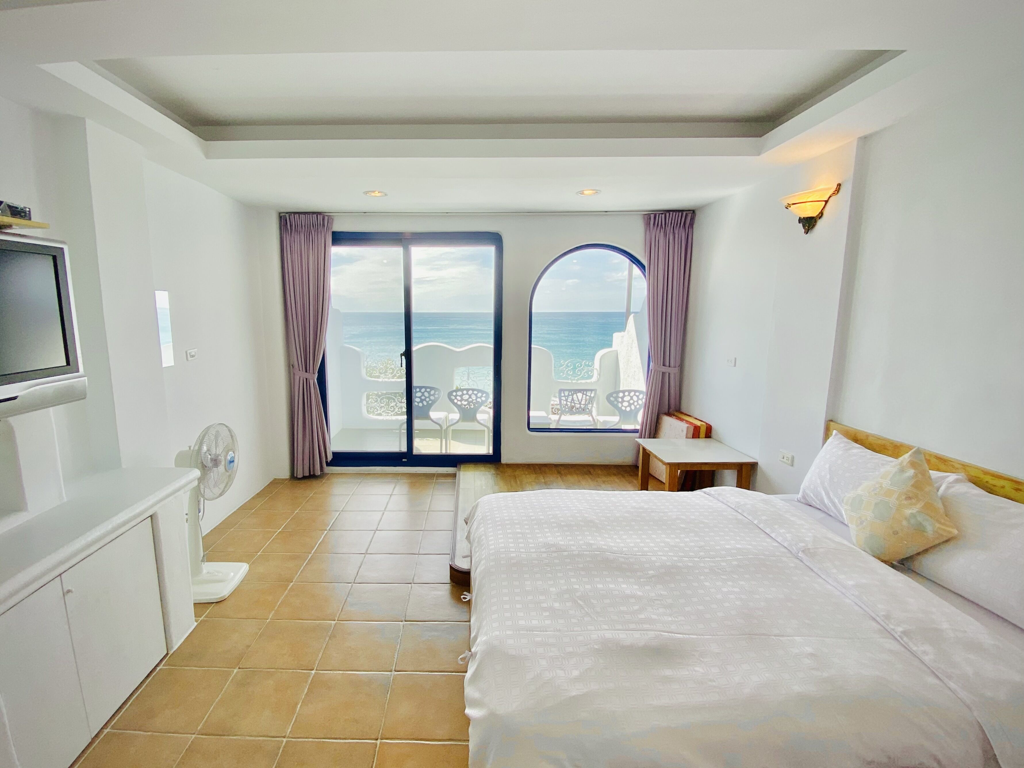 Habitación de hotel con cama, televisión, ventilador y vista al mar a través de un ventanal grande.