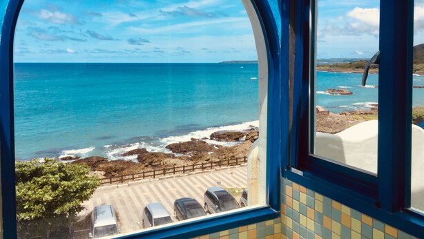 A view from a window overlooking a beach with clear blue water, rocky shoreline, and a parking area with cars. - Seya Inn (Hengchun)