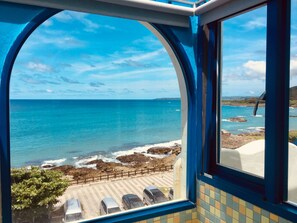 Vista desde una ventana que da a una playa con agua azul cristalina, costa rocosa y un estacionamiento con coches.