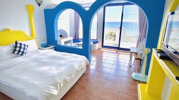 A hotel room with a bed, a desk, a chair, a TV, and a balcony with a view of the sea.