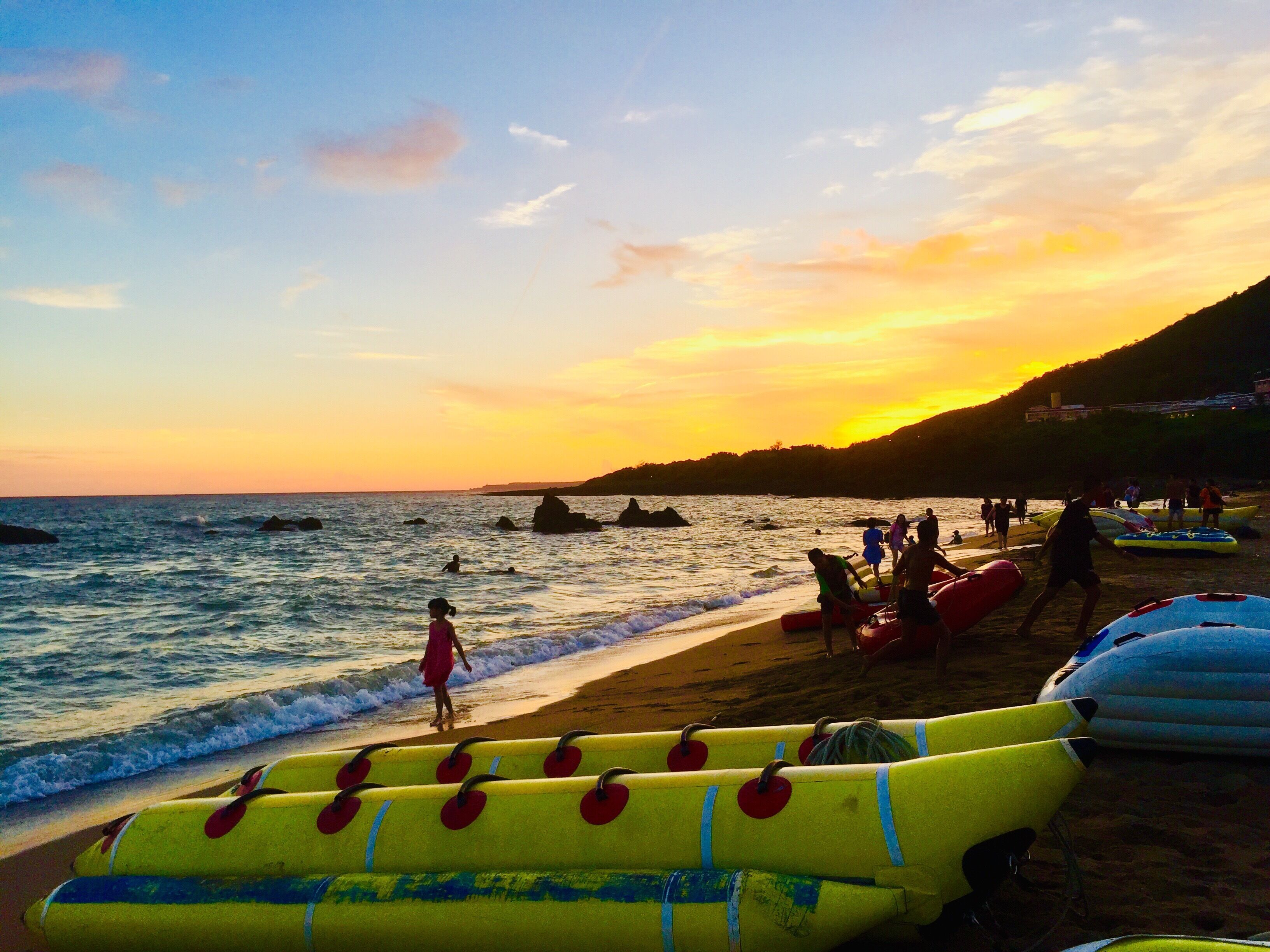 A beach with inflatable boats and people near the water during sunset.
