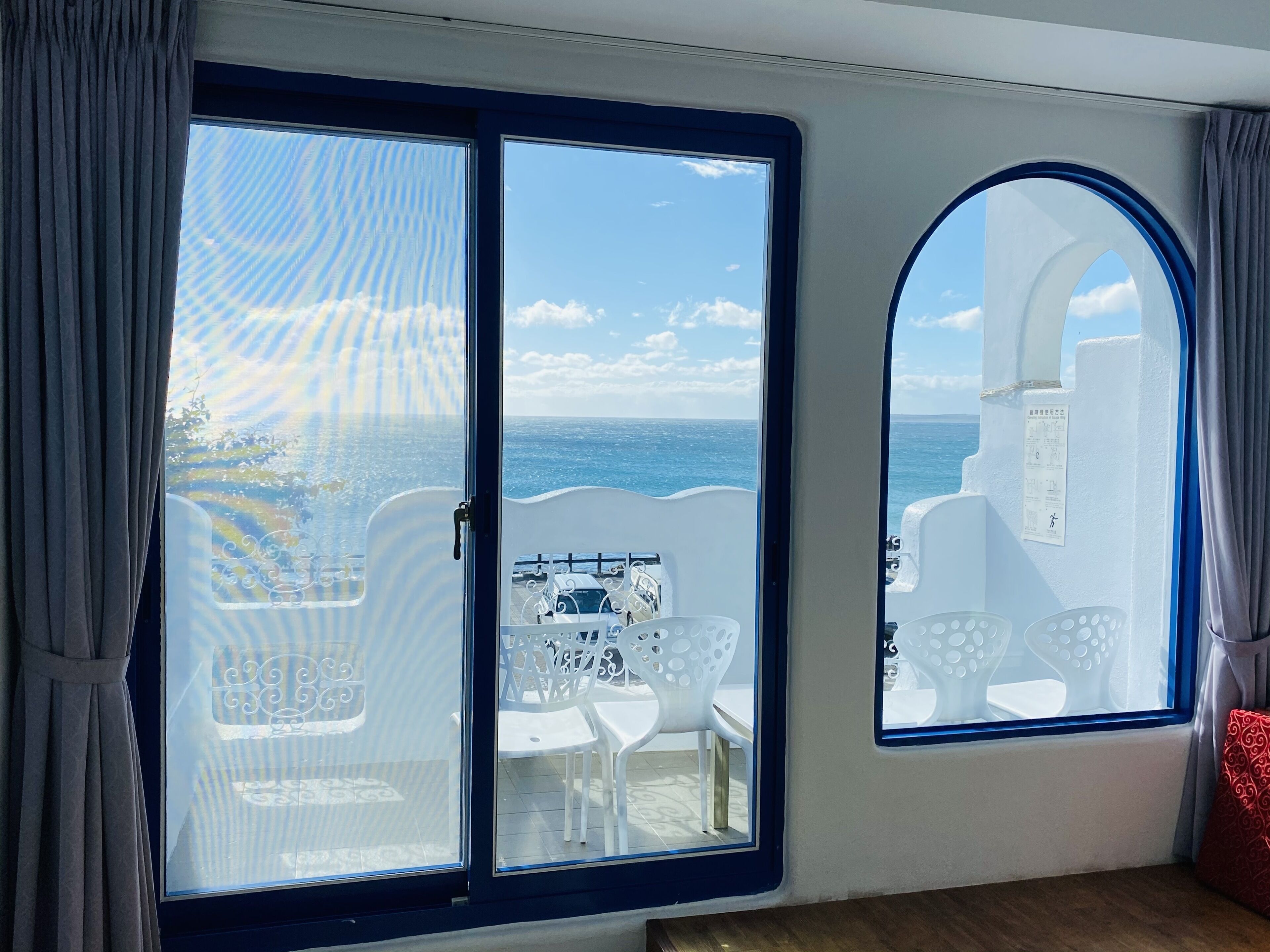 Een kamer met een grote schuifglazen deur naar een balkon met wit tuinmeubilair en uitzicht op zee.