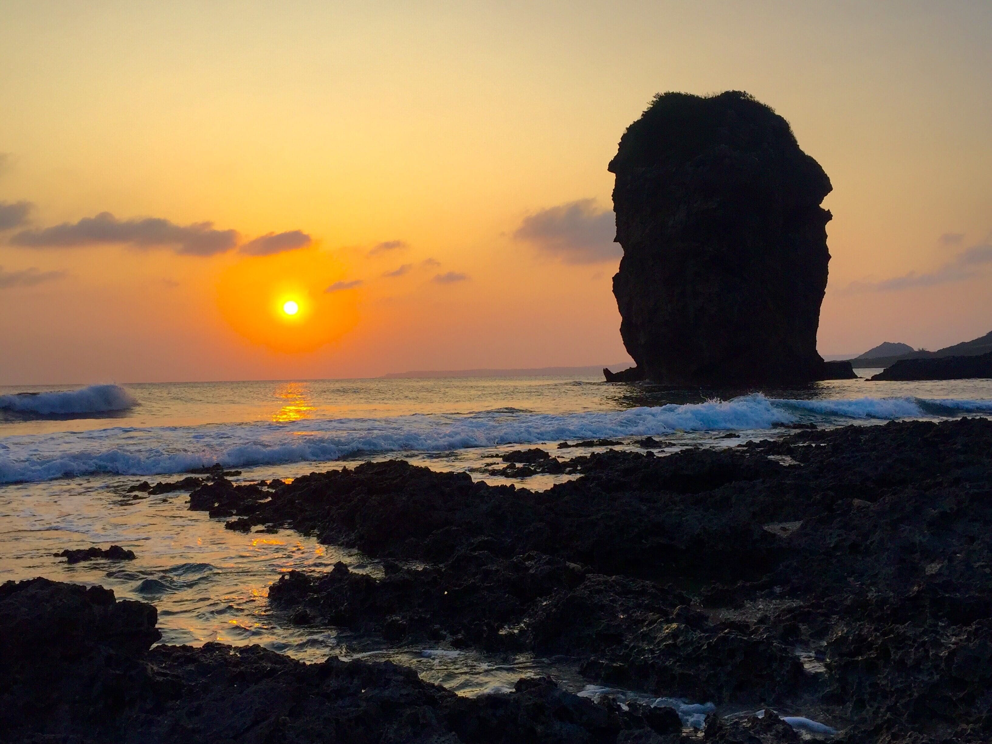 A sunset over a rocky coastline with a prominent rock formation in the center.