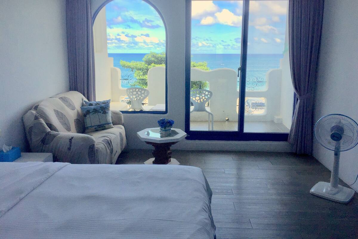 A hotel room with a bed, a sofa, a small table with a vase, a fan, and a view of the sea through a large window.