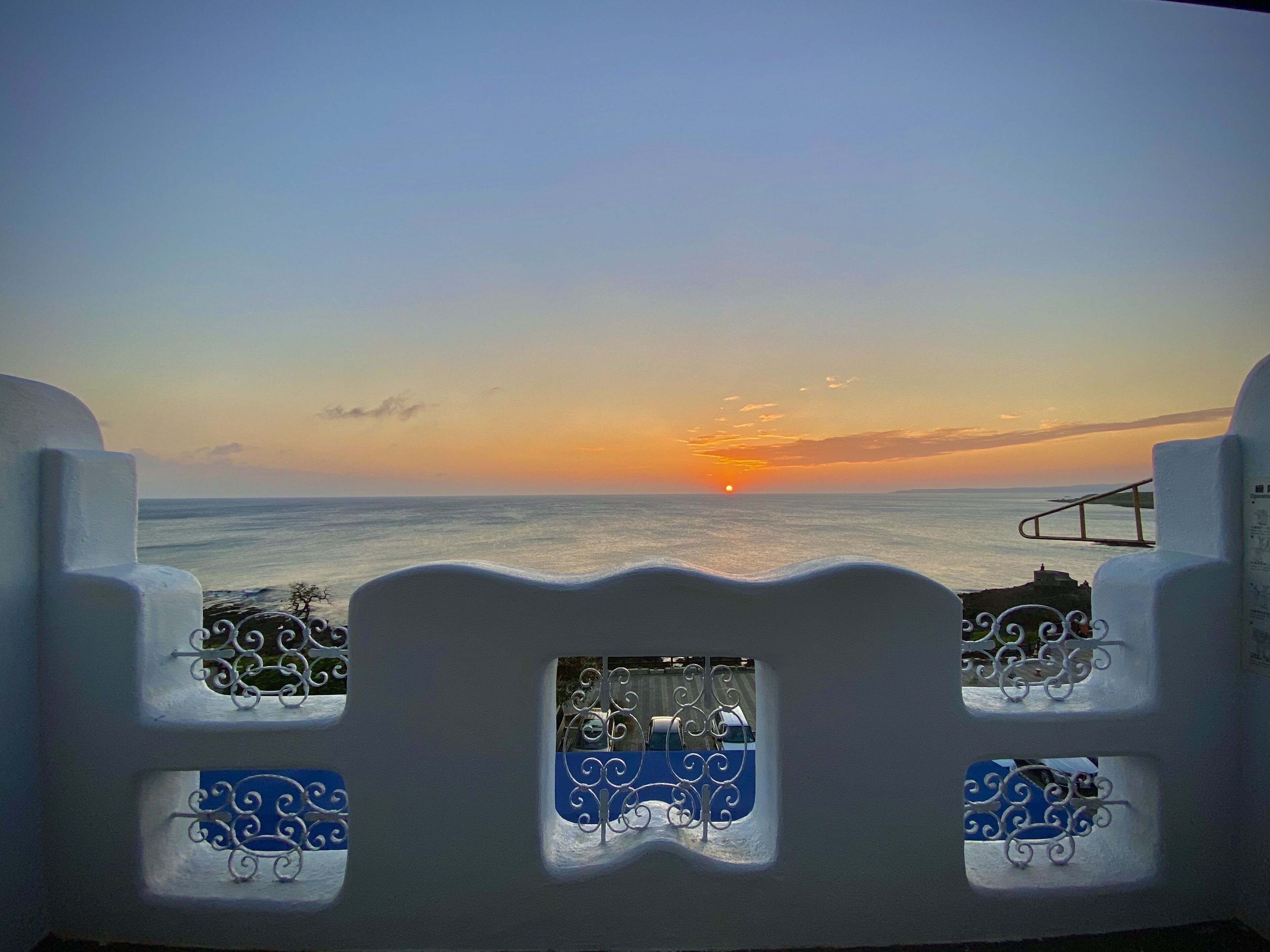 A balcony with a view of the sunset over the sea, featuring white walls and blue decorative elements.