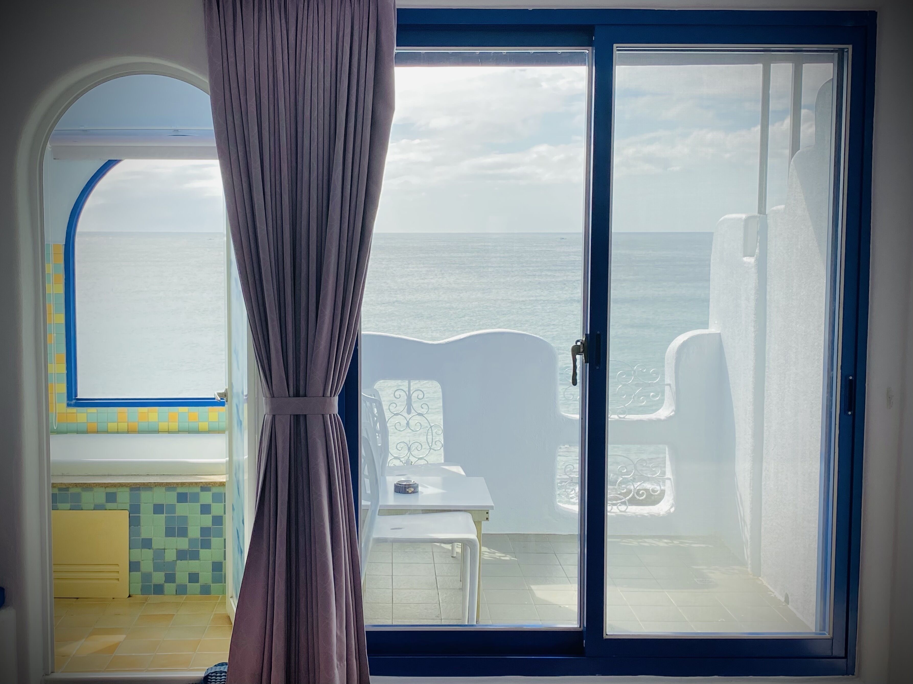 A room with a view of the sea, a sliding glass door, a small table, and a chair.