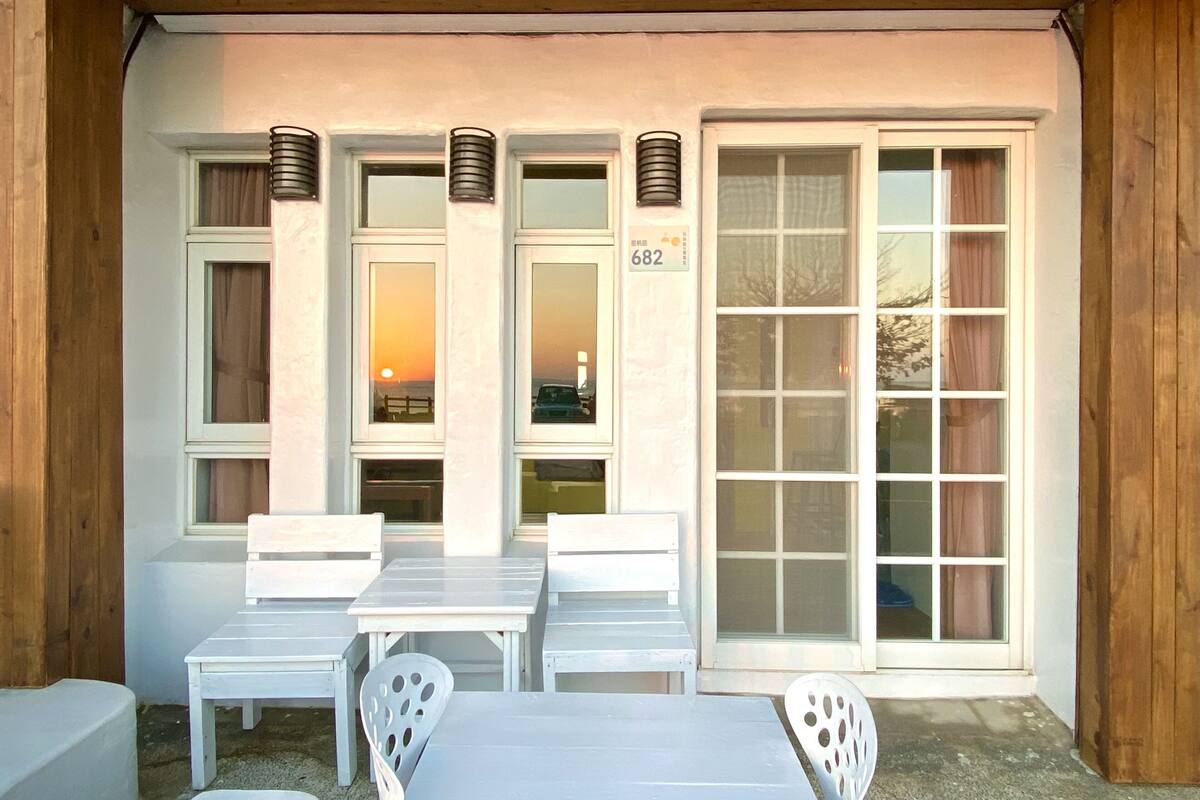 A covered outdoor seating area with white tables and chairs, a wooden ceiling, and a large sliding glass door.