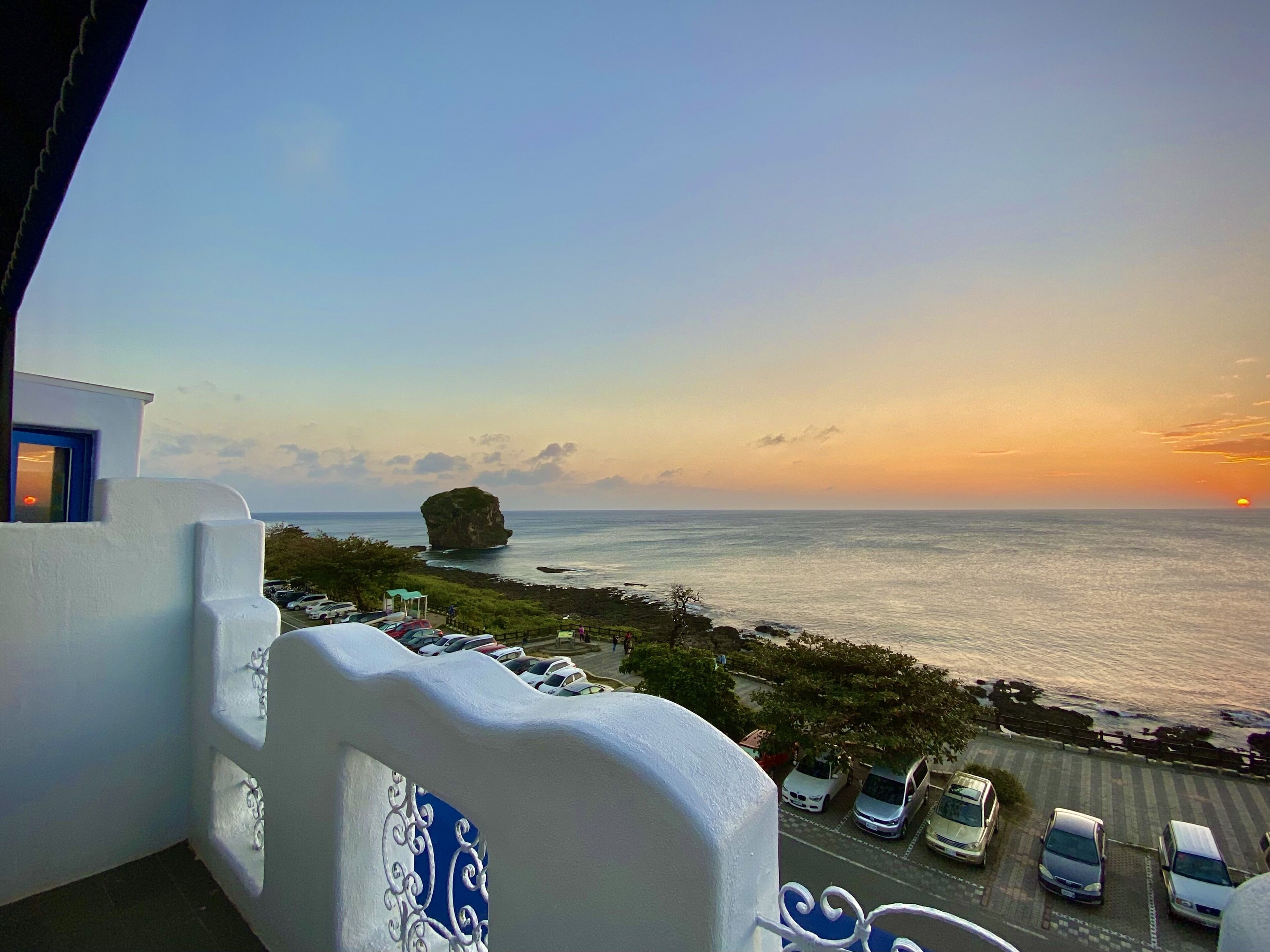 A balcony with a view of the sea, a parking area, and a building with a white facade.