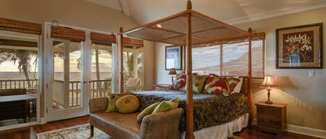 A bedroom with a four-poster bed, a sitting area with a chair and a bench, a nightstand with a lamp, and a view of the beach through large glass doors.