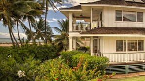 Une maison à deux étages dotée d’un balcon, entourée d’une végétation luxuriante et de palmiers, et offrant une vue sur une plage.