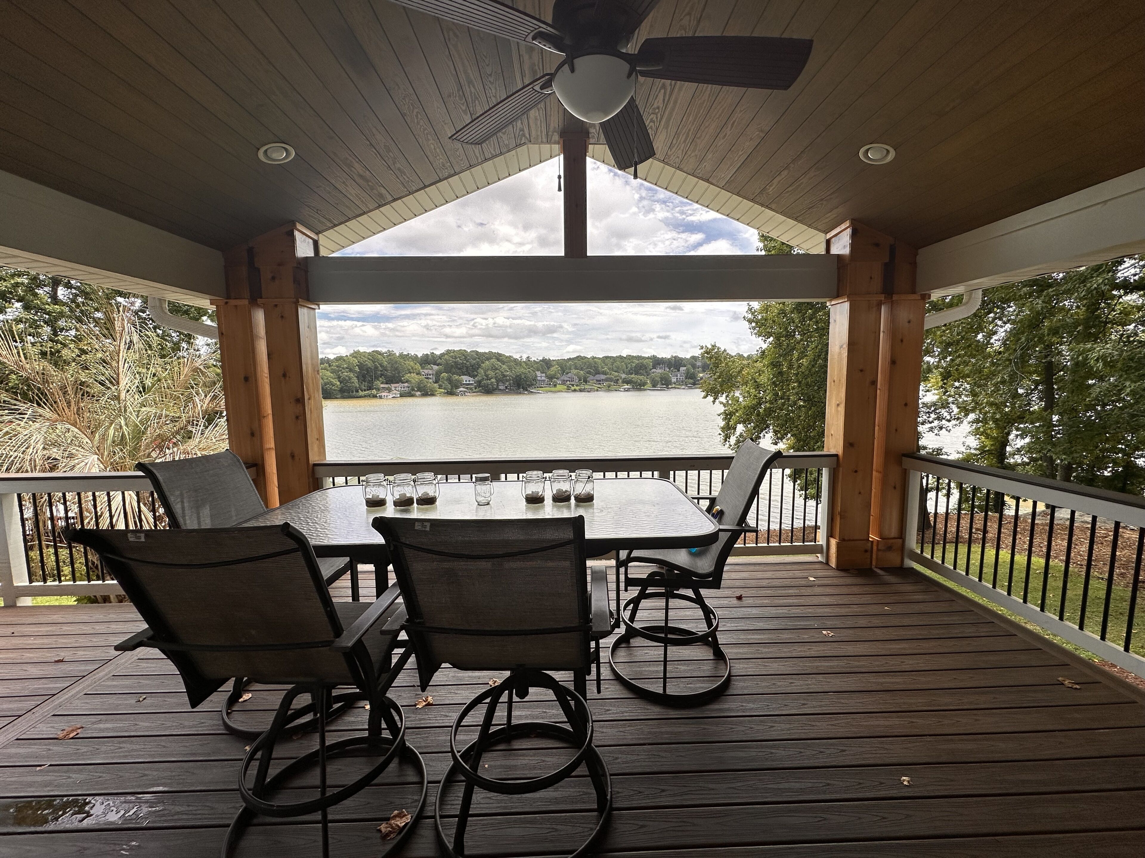 A covered outdoor deck with a table and chairs, overlooking a lake with houses in the background.