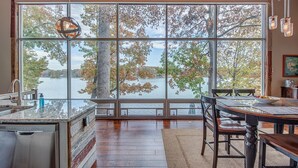 A kitchen with a large window overlooking a lake, wooden flooring, and a dining area with a table and chairs. - Firefly • Lakefront Estate • Dock + Beach + Kayaks (Belmont)
