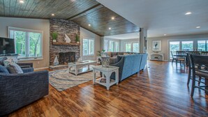A spacious living room with a stone fireplace, wooden flooring, and a large window overlooking a scenic view. - Firefly • Lakefront Estate • Dock + Beach + Kayaks (Belmont)