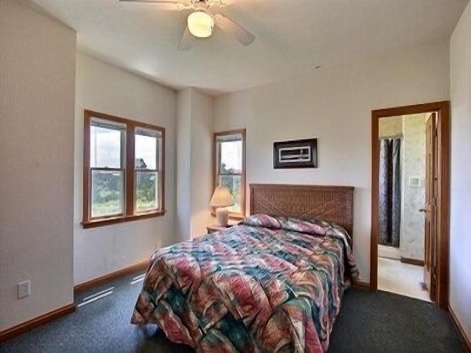 A bedroom with a bed, two windows, a ceiling fan, and a door leading to another room.
