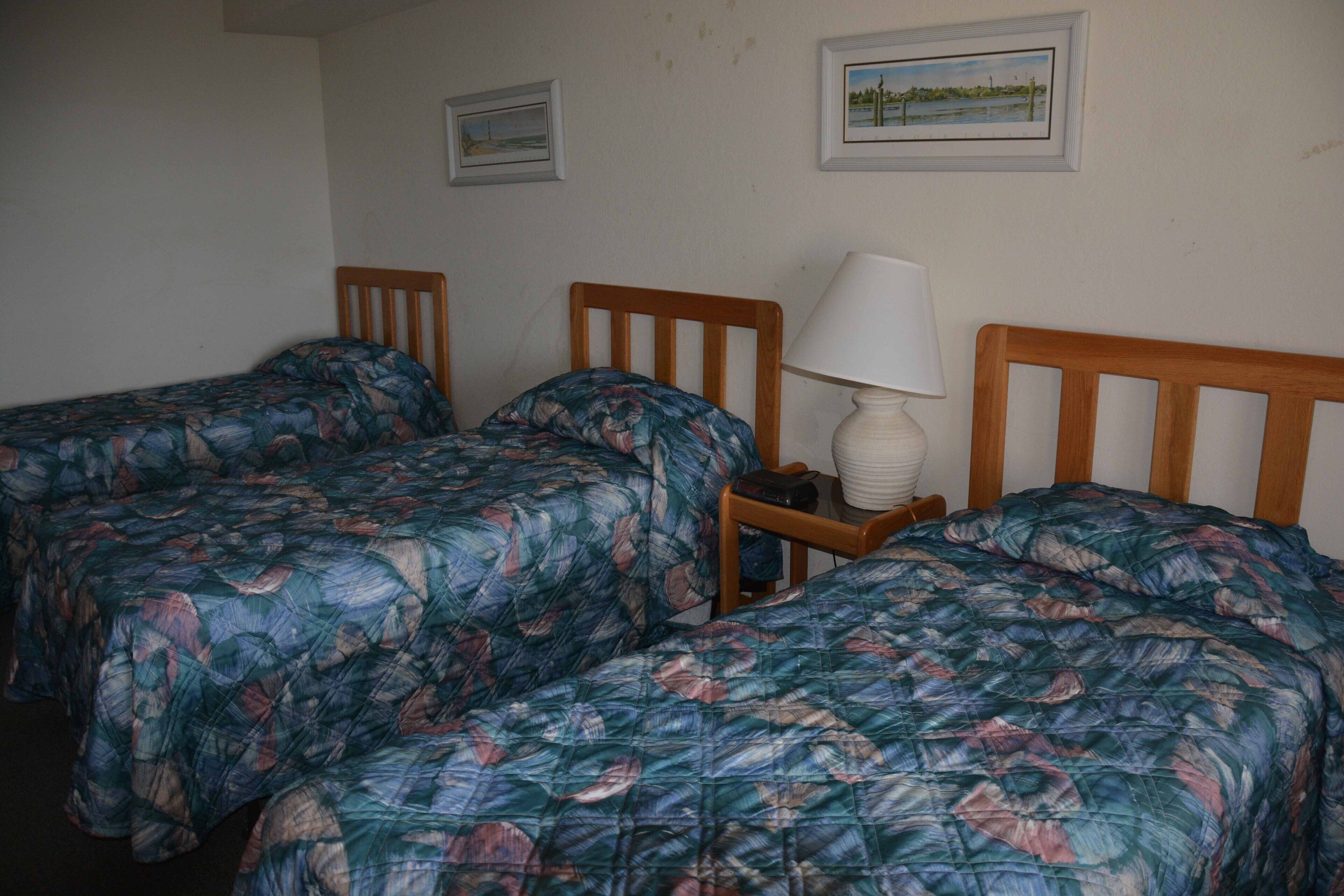 A room with two single beds, each with a wooden headboard and a floral-patterned bedspread. There is a bedside table with a lamp and a small decorative item.